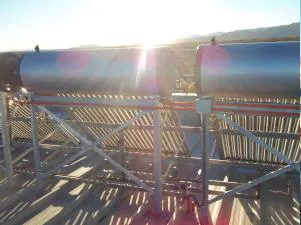 Vista trasera de calentadores solares instalados en un techo metálico al atardecer. Los tanques cilíndricos reflejan la luz del sol, mientras que las estructuras de soporte proyectan sombras en la superficie del techo.